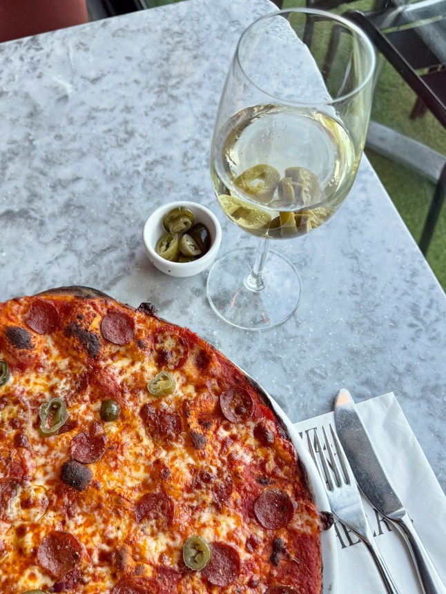Glass of white wine with a pizza next to it, and slice of jalepeno floating in the wine