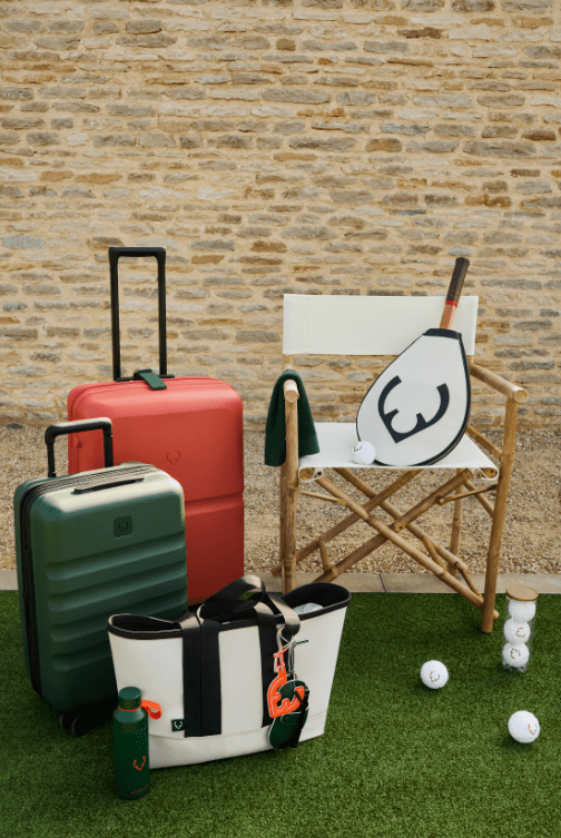 Green and red suitcase infront of each other, with a off white tote bag on the floor, and tennis racket case at an angle on a white chair