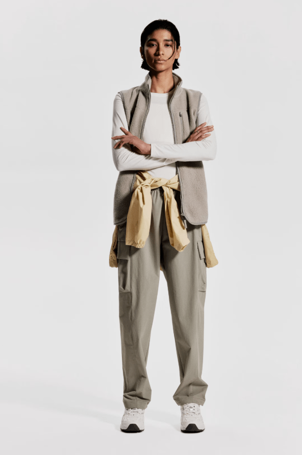 Female wears beige trousers, with jumper around the waist and gilet