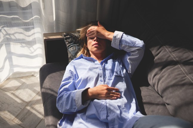 Woman exhausted and lying on couch. Hand on forehead, eyes closed. Indoors with natural light near a window with curtains.
