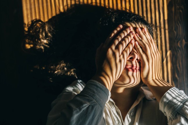 A depressed woman lies on wooden floor and closes her face with hand, striped from blinds on face. Top view.