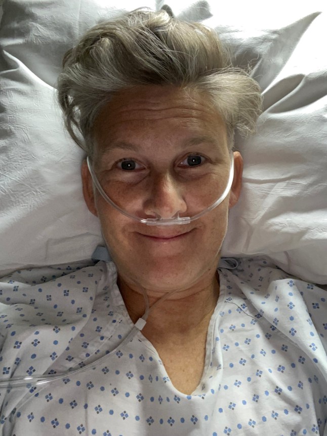 Kit takes a selfie in bed, wearing a hospital gown