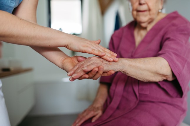 Home caregiver helping elderly woman with skincare in bathroom, applying moisturise lotion on hands, skin. Senior woman at home with nurse, close up.