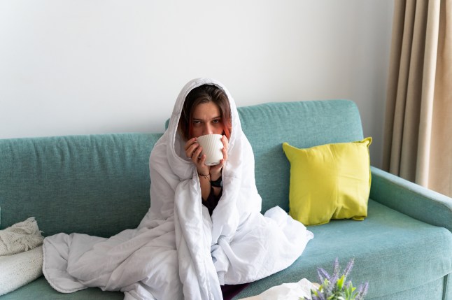 Woman bundled up under a blanket, drinking hot tea on sofa at home