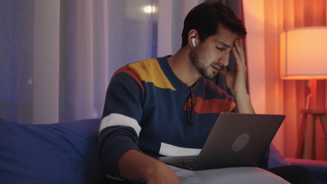 A Man looking stressed holding his head in frustration while working on his laptop at home on the sofa