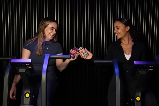 Two people on exercise bikes with a can of Hip Pop each cheersing