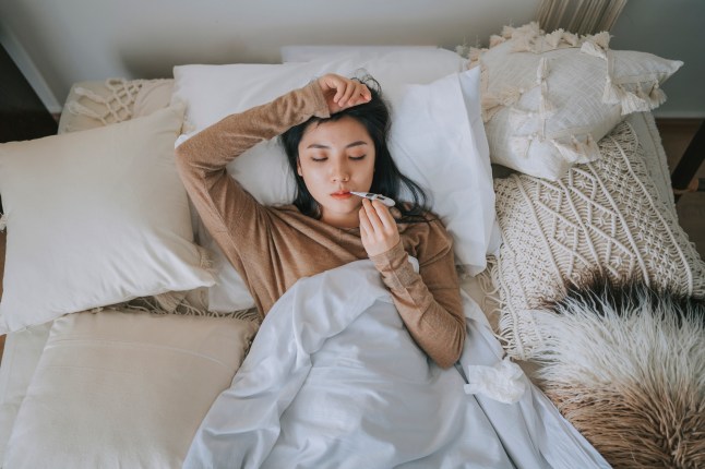 Woman lying on bed covered with blanket having cold and flu measuring her body temperature with a digital thermometer.