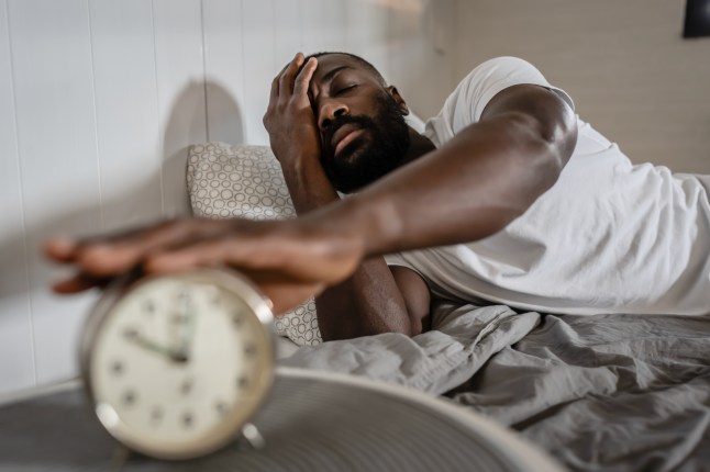 A tired man in a white t-shirt lies in bed, reaching to turn off his alarm clock with a sleepy expression. He appears exhausted, struggling to wake up in the morning.