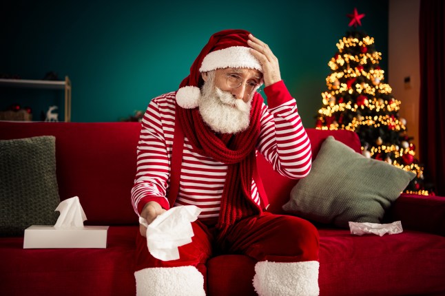 Elderly man dressed as Santa sitting on a sofa with a tissue, amidst festive Christmas decor and tree