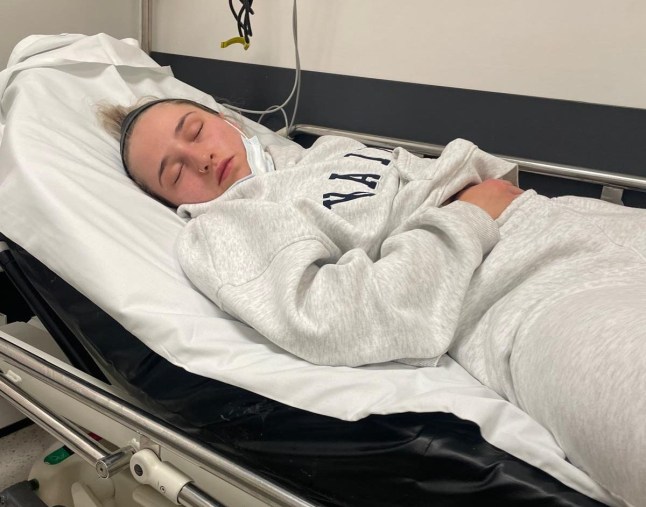 Alice Cooper in a grey tracksuit laying on a hospital bed and holding her stomach