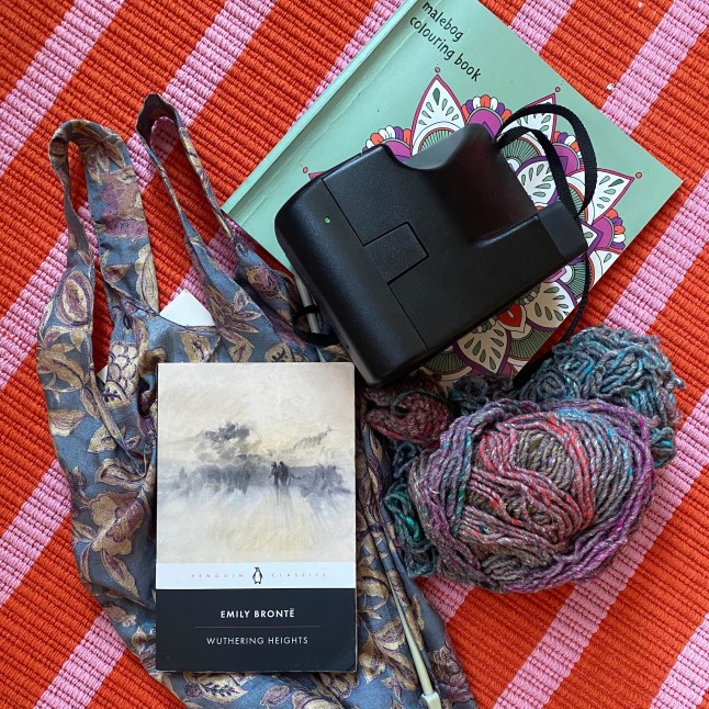 The contents of Eleanor Noyce's analogue bag concept, featuring a colouring book, Polaroid camera, silk tote bag, a copy of Wuthering Heights by Emily Bronte, and some knitting.